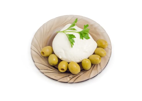 Ball of the fresh soaked mozzarella cheese with twig of parsley and several green olives on the glass saucer on a light background
