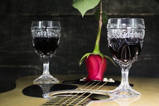 Glass Of Red Wine Resting on Acoustic Guitar With Red Rose and another Glass in The Background 