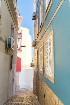 Stairs made of portuguese sidewalk rocks leading a way down to the entrance of a beautiful blue house with white metal shutters. on the other side a older facade of another house. Blue sky in the background.