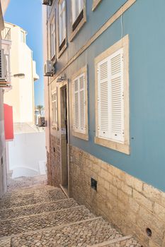 Stairs made of portuguese sidewalk rocks leading a way down to the entrance of a beautiful blue house with white metal shutters. Blue sky in the background.