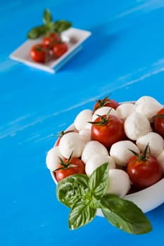 Closeup of fresh mozzarella, cherry tomatoes and basil in a white bowl over a blue wooden background