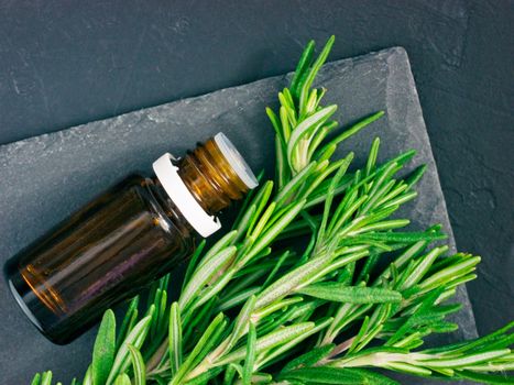 Rosemary essential oil in dark glass bottle and fresh rosemary on dark background. Top view or flat lay
