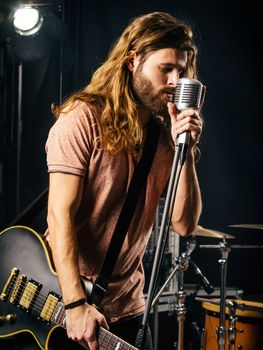 Photo of a young man with long hair and beard singing and playing electric guitar on stage.