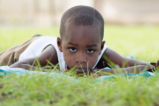 This child lying on a piece of cloth , looking at the camera , sits in the shade of a tree on the lawn.