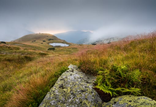 Foggy Mountain Landscape with a Tarn and Rocks in Foreground at Sunset