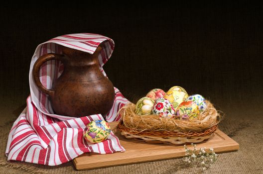 Decoupage Easter eggs in a basket and a jug under the towel, the dark background is burlap. Selective focus.