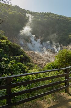 A small fence in the forground frames this live volcano smoking at Soufriere, Saint Lucia. The smoke rolls up the tropical rainforest hill.