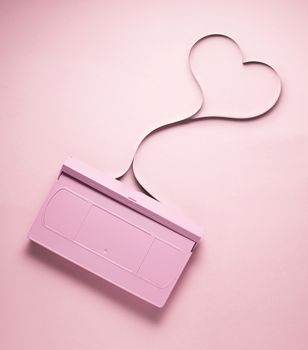 Creative love concept photo of painted VHS cassette with tape in the shape of heart on pink  background.