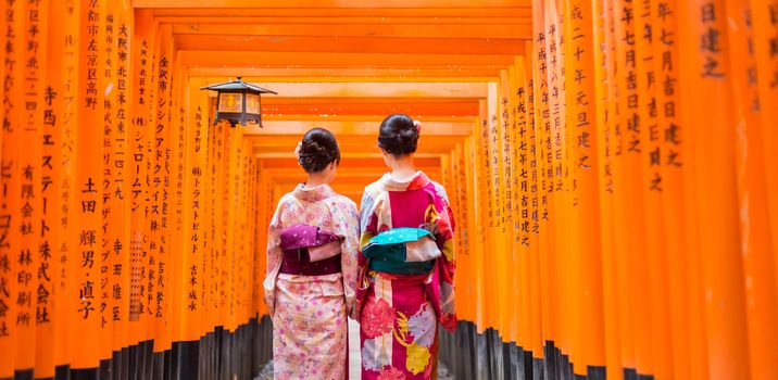 Two geishas among red wooden Tori Gate at Fushimi Inari Shrine in Kyoto, Japan. Selective focus on women wearing traditional japanese kimono.