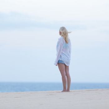 Meditative, sensual blonde woman wearing white loose casual shirt on vacations looking at horizon at dusk.