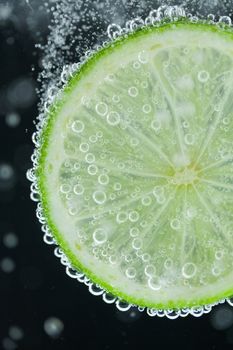 Close-up of single green lime slice falling into carbonated water with bubbles on black background. Refresher drink concept