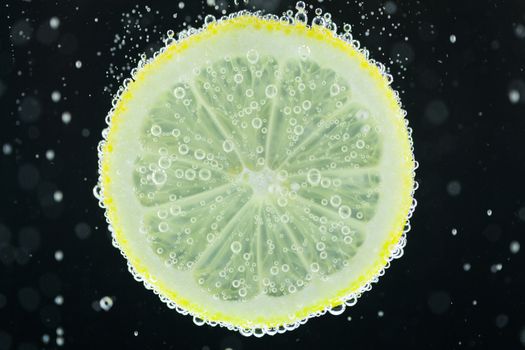 Close-up view of lemon slice under carbonated water with bubbles, refresher concept on black background