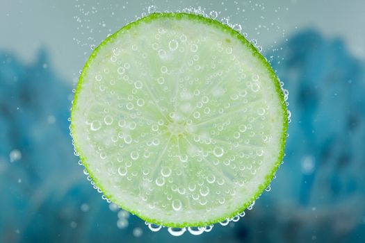 Close-up of lime green slice on blue background diving inside carbonated water with bubbles