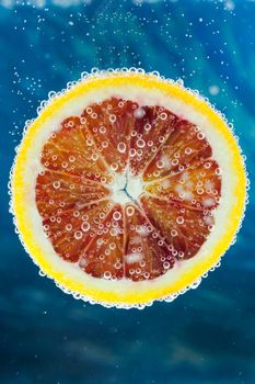 Close-up of slice of citrus blood orange dropped into carbonated water with bubbles on blue background