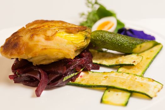 Delicious gourmet plate featuring biscuit sandwich with beets, zucchini strips and pansy garnish over white background