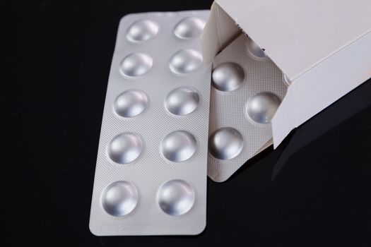 Silver blister pack of small pills together with the a box with additional packs showing through the open lid over a black background in a healthcare concept