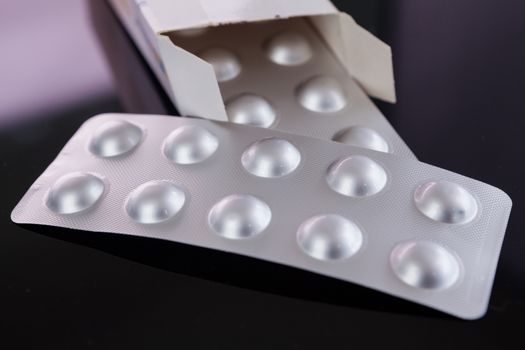 Silver blister pack of small pills together with the a box with additional packs showing through the open lid over a black background in a healthcare concept