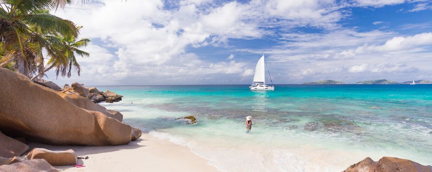 Woman wearing long floral summer dress and beach hat on Anse Patates beach on La Digue Island, Seychelles. Summer vacations on picture perfect tropical beach concept.