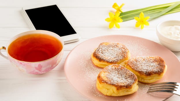 Cheese pancake with powdered sugar, black tea, flowers and smartphone on a white wooden table.