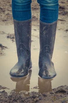 A Child's Legs In Black Rubber Boots Standing In A Muddy Autumn Puddle