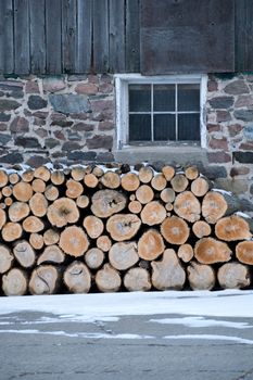 Neatly stacked firewood by an old Ontario Barn.  Barn is barnboards, field stone and has windows too. Snow on the ground.