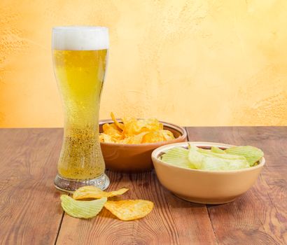 Beer glassware with the lager beer, two different kinds of the potato chip flavored paprika and wasabi in ceramic bowls on an old wooden table on a background of an yellow wall
