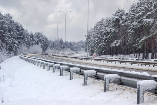 Part of the motorway with cars, traffic barriers, lampposts and pine forest covered snow on both sides of road after snowfall in cloudy day
