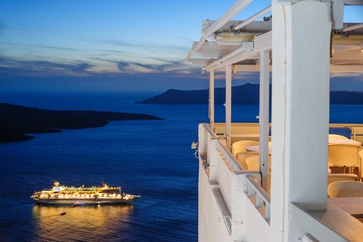Enlighted cruise ship just after sunset near Fira town at Santorini island, Greece, empty outdoor cafe to the right and caldera sea at background