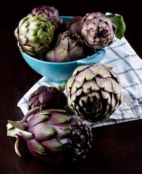 Stack of Perfect Raw Artichokes in Blue Colander on Checkered Napkin closeup on Dark Wooden background