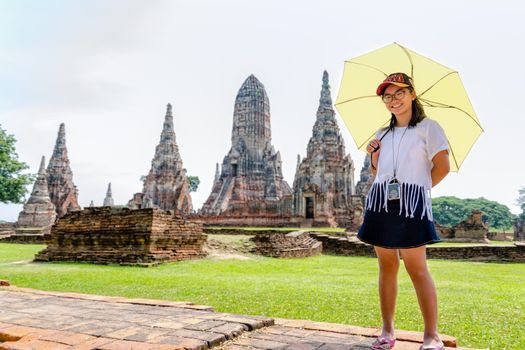 Tourist cute teenage girl wearing glasses with a camera neck, looking and smiled happily holding the umbrella on Wat Chaiwatthanaram temple background in Ayutthaya Historical Park, Thailand