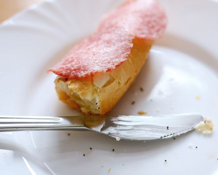 Leftover of salami bread roll with missing bite and lying knife on a white plate.