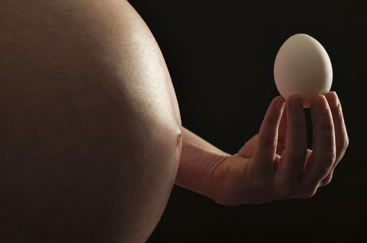 A close-up of pregnant female abdomen with her hand holding an egg on black background.