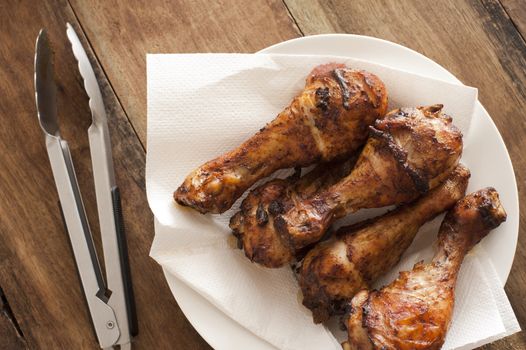Four delicious marinated grilled chicken legs arranged in a line on paper on a plate with tongs alongside, overhead view