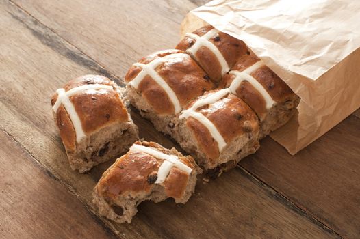 Hot Cross Buns for Easter in a brown paper packet spilling out onto a wooden table with one bun having a missing bite