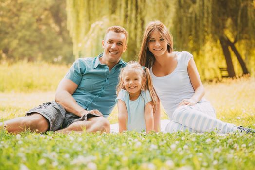 Beautiful smiling family sitting on the grass in the park and looking at the camera.