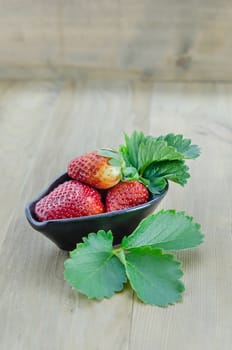 fresh strawberry fruits in black bowl with green leaves over wooden background