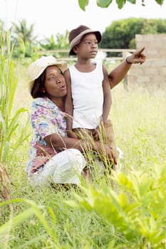 This young woman squatting shows something to her child in the bush.