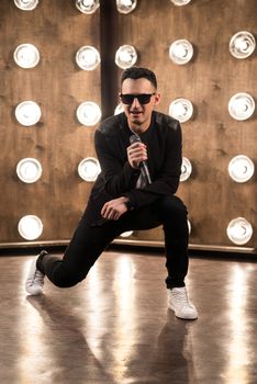 Male singer of rock or pop music dressed in black and sunglasses with microphone performs on scene with lightening projectors on background