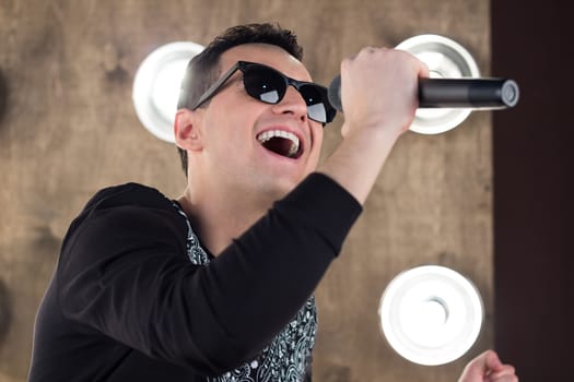 Male singer of rock or pop music dressed in black and sunglasses with microphone performs on scene with lightening projectors on background