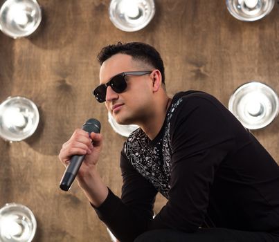 Male singer of rock or pop music dressed in black and sunglasses with microphone performs on scene with lightening projectors on background