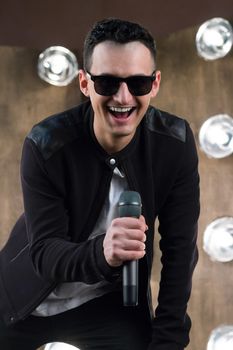 Male singer of rock or pop music dressed in black and sunglasses with microphone performs on scene with lightening projectors on background