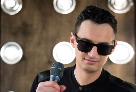 Male singer of rock or pop music dressed in black and sunglasses with microphone performs on scene with lightening projectors on background