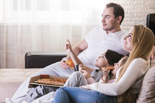 Young parents and their children are watching TV, eating pizza and smiling while sitting on couch at home