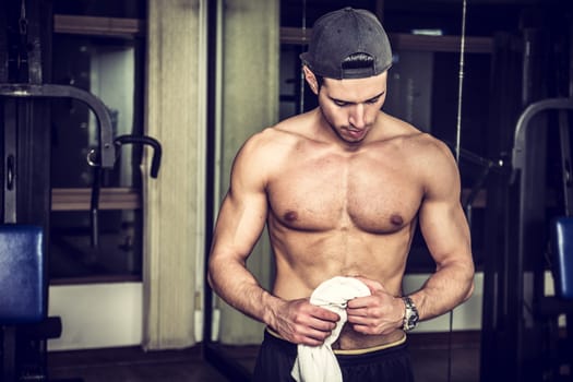 Muscular, shirtless young man resting in gym during workout, showing muscular torso, pecs and abs, holding a towel in his hands