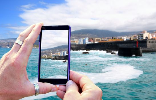 View over the mobile phone display during shooting of Tenerife coast. Holding the mobile phone in hands and taking a photo, focused on mobile phone screen.