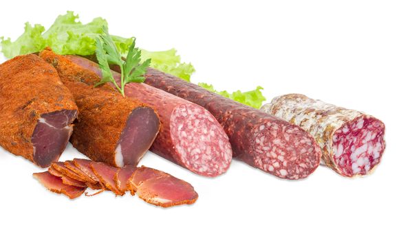 Fragment of the partly sliced two pieces of the dried pork tenderloin, two different dry smoked sausage with parsley twig and salami on the lettuce closeup on a light background  
