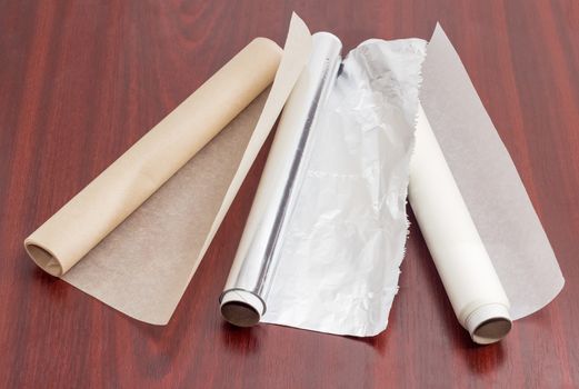 Two rolls of the various parchment paper and one roll of the aluminum foil for household use on a dark red wooden surface
