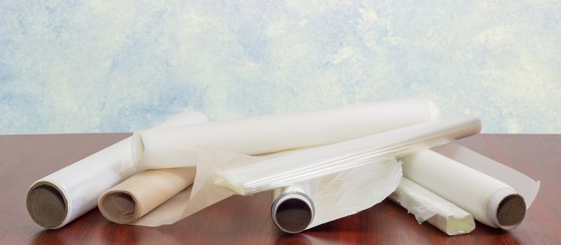 Several rolls of the plastic oven bags, plastic food wrap, aluminum foil and various parchment paper for household use closeup on a dark red wooden table 
