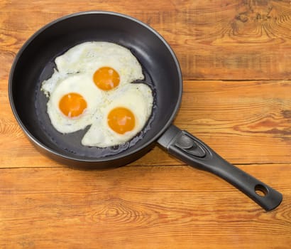 Three freshly fried eggs prepared with unbroken yolk in the frying pan on a surface of an old wooden planks
