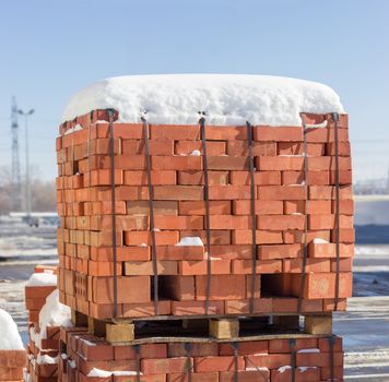 Wooden pallet of the red bricks covered snow on an another such pallet on a outdoor warehouse in winter sunny day
